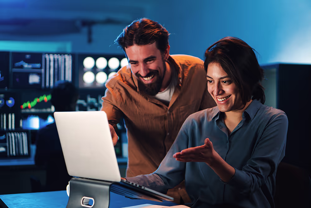 Smiling man and woman collaborating enthusiastically over an open laptop in a tech-focused office.