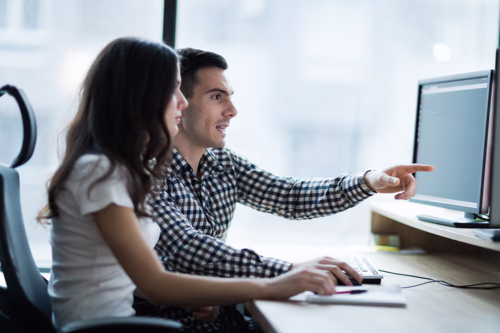 Two business people working together at a desk, with one pointing at a computer screen.