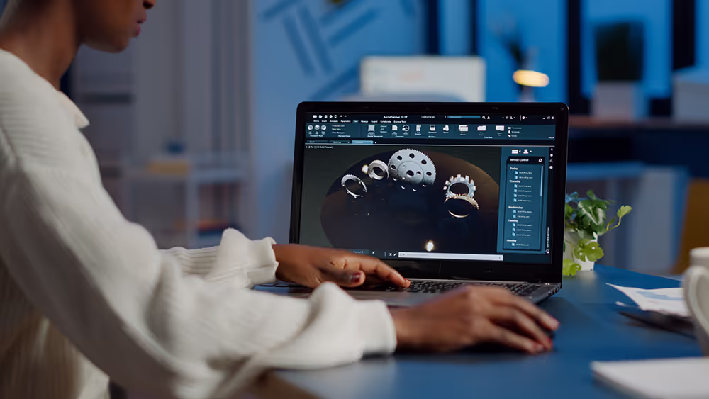 Person working on a laptop with 3D mechanical part designs on the screen in an office setting.