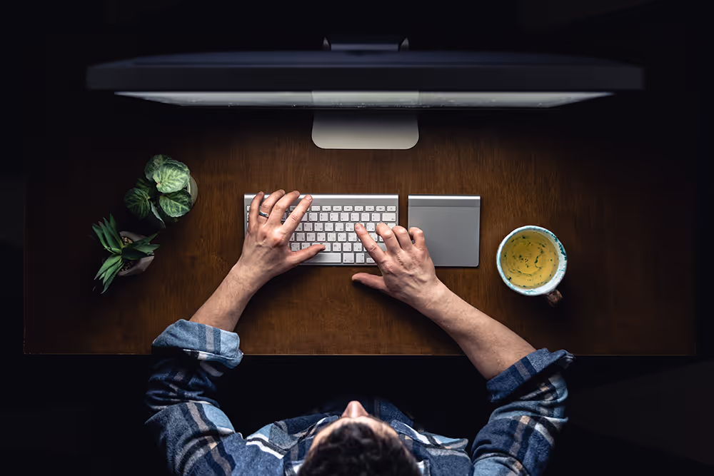 Top view of a person typing on a wireless keyboard at a wooden desk with two small plants on the left and a cup of coffee on the right.